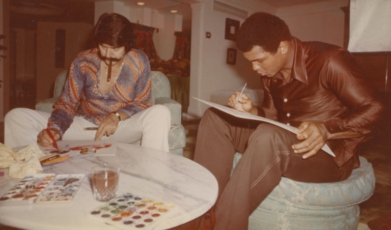 Vintage 1970s photo of Muhammad Ali and a man with a mustache seated painting in watercolors
