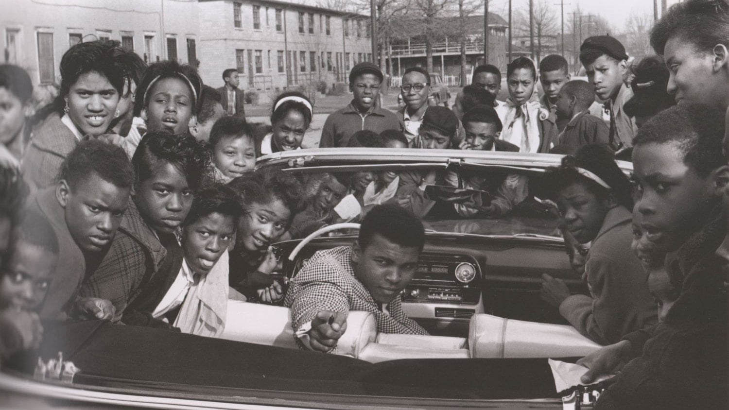 Black and white photo of Muhammad Ali as a young man sitting in a convertible and dramatically pointing while surrounded by a crowd of people around the car