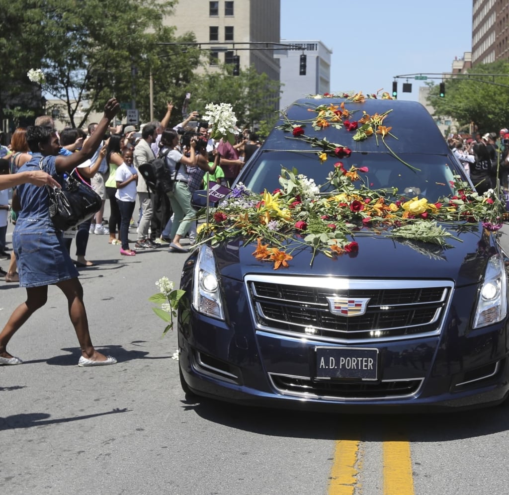 Hearse covered in flowers drives down street with crowd throwing flowers onto the car
