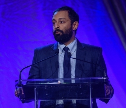 Man wearing dark jacket with blue shirt and tie speaks at lecturn