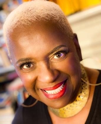 Woman with short blonde hair and bright red lipstick looking at the camera wearing a large gold necklace and black top