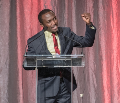 Man with black jacket, light green shirt and red tie raises fist while at lecturn