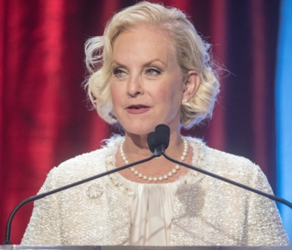 Woman wearing white blouse with white pearl necklace speaks at lecturn