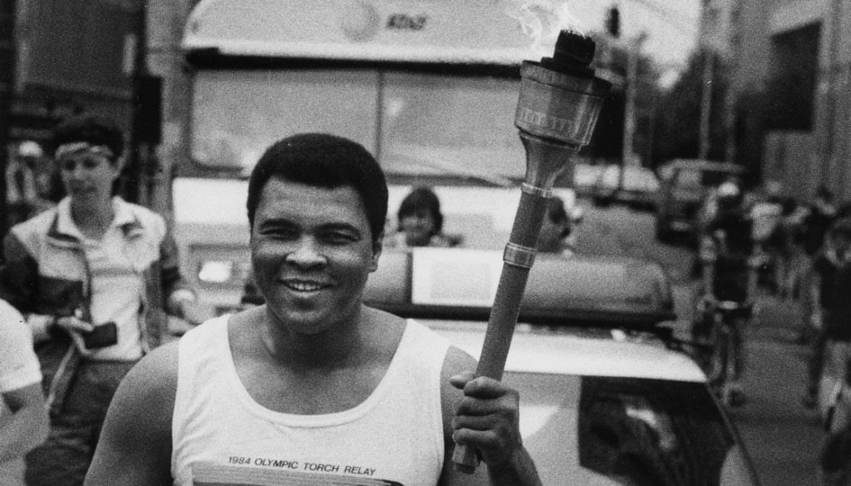 Black and white photo of Muhammad Ali running with the Olympic Torch in the middle of a street