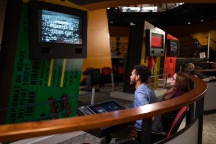 Two people in an exhibit sitting and watching an old black and white Muhammad Ali fight