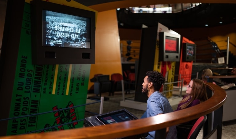 Two people in an exhibit sitting and watching an old black and white Muhammad Ali fight