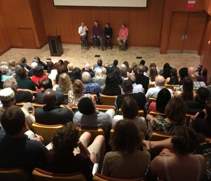 A group of people sitting in an auditorium while four presenters speak