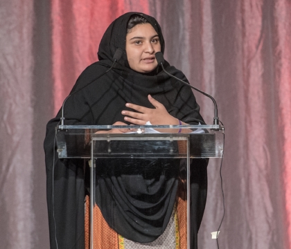 Woman wearing hijab and red and white dress speaks at lecturn holding hands at heart