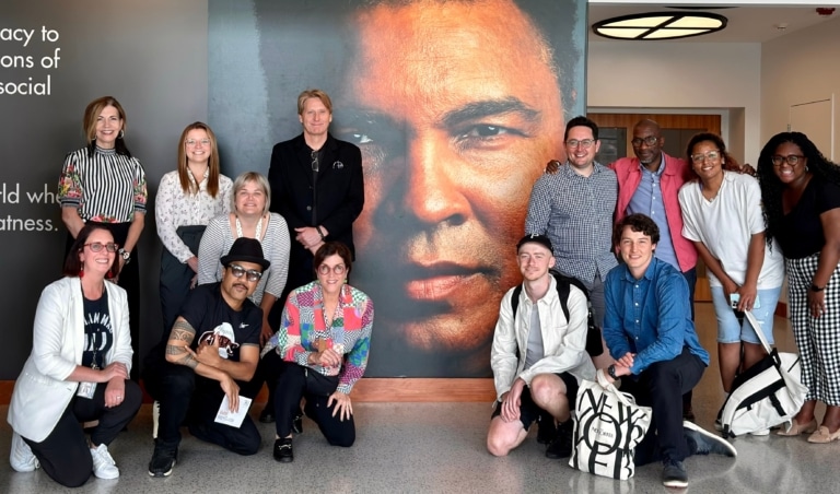 Group of people kneeling and standing in front of a large wall with a photo of Muhammad Ali's face