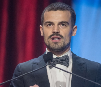 Man wearing black jacket, white shirt and black bowtie speaks at lecturn