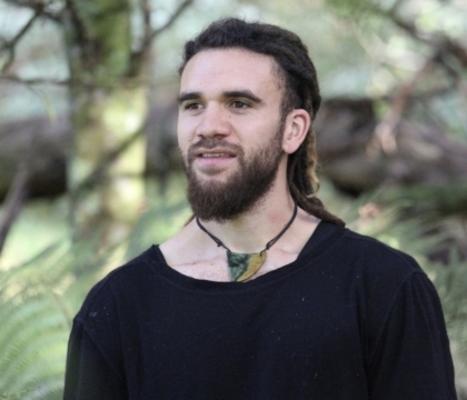 Man with beard wearing black shirt and necklace posing in front of greenery