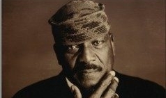 Jim Brown, wearing black jacket, white shirt and kufi hat poses with hand to his chin