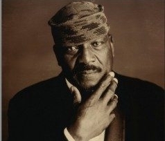Jim Brown, wearing black jacket, white shirt and kufi hat poses with hand to his chin