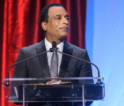 Man with black jacket, gray tie and light-colored shirt looks off-camera while at lecturn