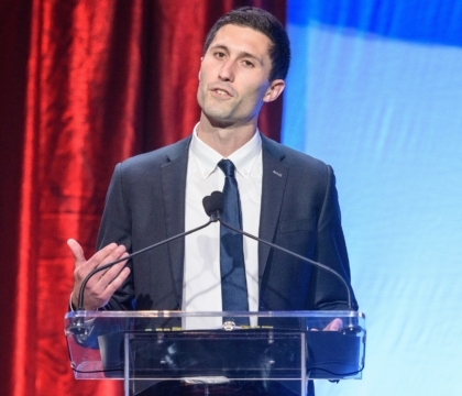 Man in black jacket, white shirt and dark blue tie speaks at lecturn