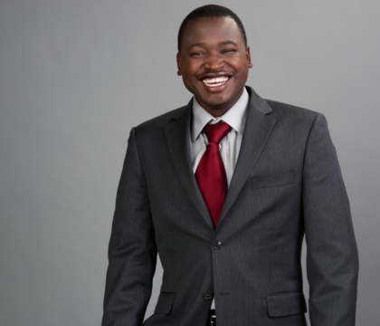 Man with black jacket, gray shirt and red tie smiling in front of gray background