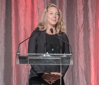 Woman wearing black dress and black jacket speaks at lecturn