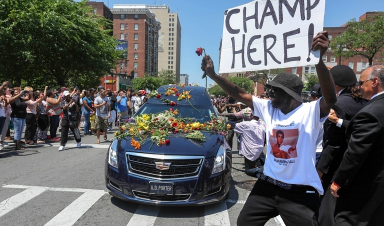Funeral hearse, convered in flowers, drives down a street surrounded by crowds of people