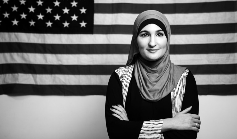 Black and white portrait photo of a woman in a headscarf standing in front of an American flag