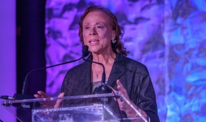 Lonnie Ali speaking on a stage while standing at a clear glass podium in front of a purple background