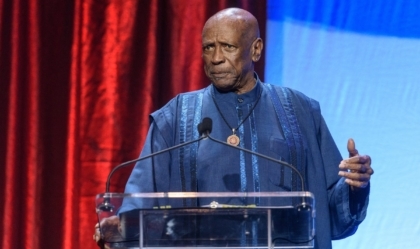 Man with blue outfit wearing necklace speaks at lecturn