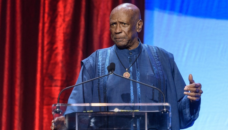 Man with blue outfit wearing necklace speaks at lecturn