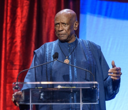 Man with blue outfit wearing necklace speaks at lecturn