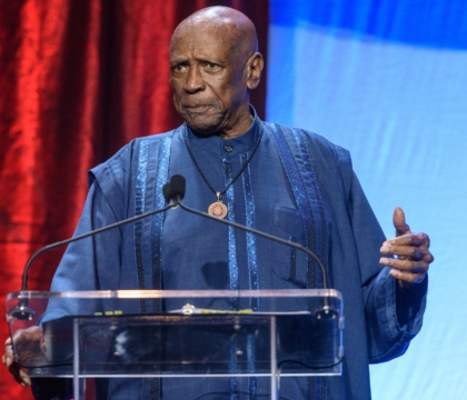 Man with blue outfit wearing necklace speaks at lecturn