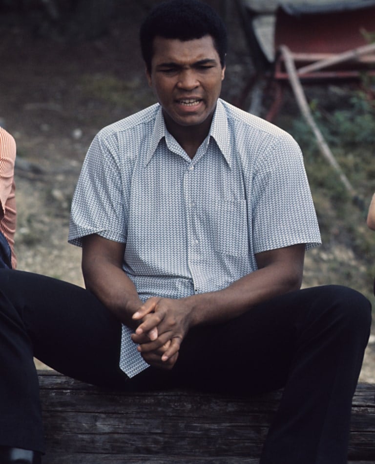 Color photo of Muhammad Ali sitting on wooden logs outside Deer Lake