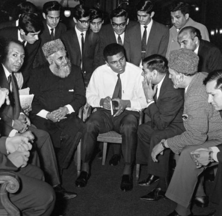 Muhammad Ali seated in a group discussion during an overseas visit, holding a book and listening intently.
