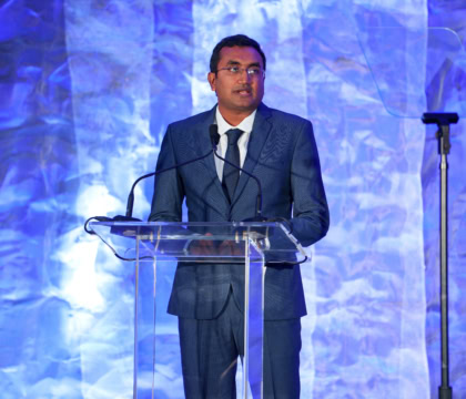 Man in blue suit and blue tie with short hair and glasses speaks at podium