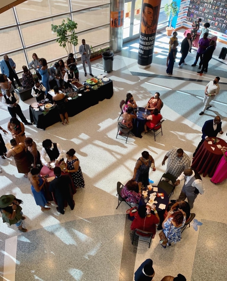 Room with a large glass wall and terrazzo flooring set up for an event with a buffet, small tables, and people standing and sitting