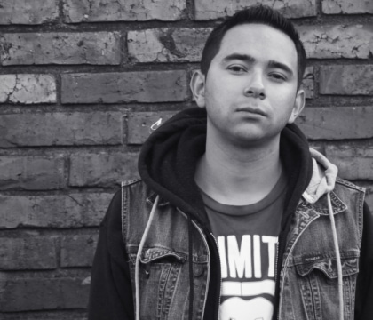 Black and white photo of man with denim vest and t-shirt posing in front of brick wall