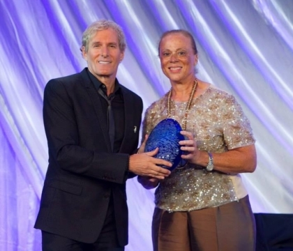 Michael Bolton, wearing black jacket, black shirt and black tie, posing with Lonnie Ali and award