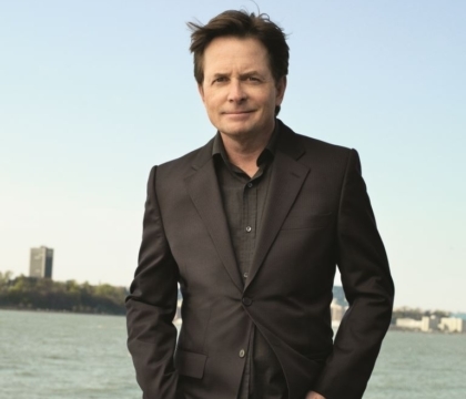 Michael J. Fox posing with black suit and black shirt in front of body of water