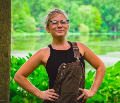 Woman with glasses wearing black shirt and overalls posing in front of greenery