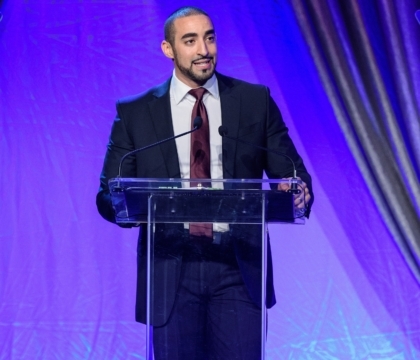 Man wearing black jacket and dark purple tie speaks at lecturn