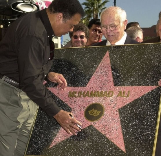 Older man holding up Hollywood Walk of Fame star while 168一分钟极速赛车官网预测最新结果-168极速一分钟赛果查询网-168官网一分钟极速赛车开奖记录 in button up shirt and gray pants signs with marker