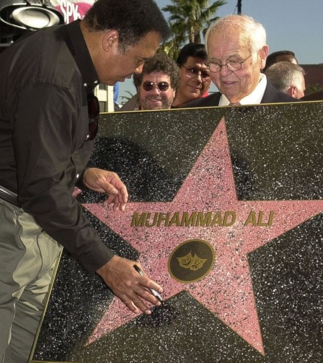 Older man holding up Hollywood Walk of Fame star while Muhammad Ali in button up shirt and gray pants signs with marker