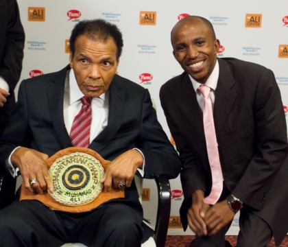 Man with black jacket, white shirt and pink tie posing with Muhammad Ali, holding a Humanitarian Champ belt