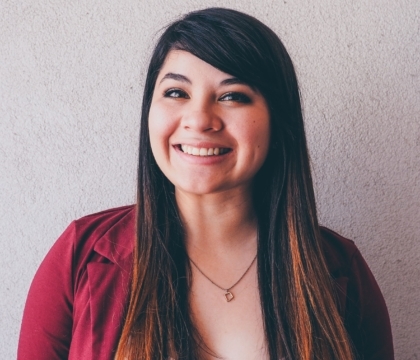 Woman wearing red jacket and necklace smiles for camera in front of gray wall