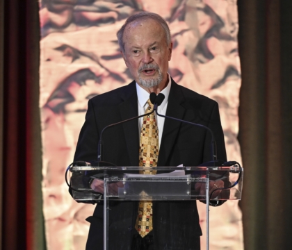 Man in black suit and brown-patterned tie speaking at lecturn