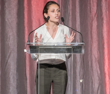 Woman wearing white blouse and dark pants speaks at lecturn with hands raised