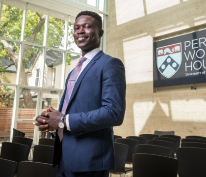 Man wearing blue suit with purple tie posing off center with sign saying "Perry World House University of Pennsylvania"