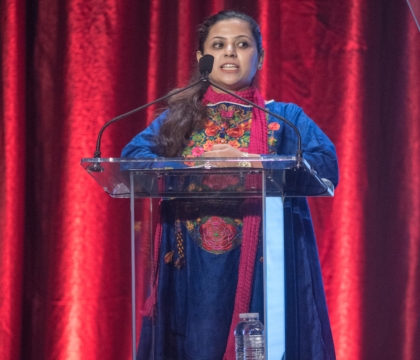 Woman wearing blue floral top with red scarf speaks at lecturn