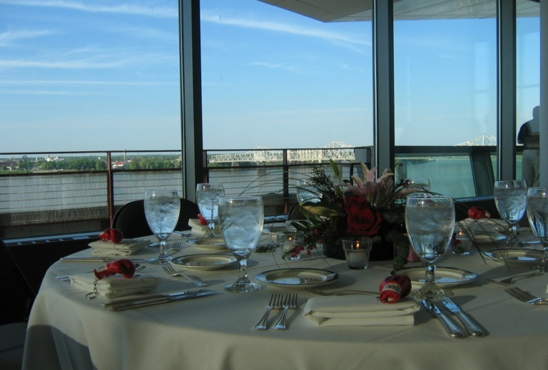 Event space with a large round table, place settings, and glasses of ice water situated in front of a large window with a view of a river and a bridge
