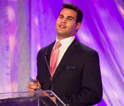 Man wearing black jacket, white shirt and pink patterned tie speaking at lecturn