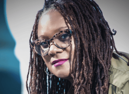 A woman with tortoiseshell glasses, braids, and purple lipstick wearing a green jacket