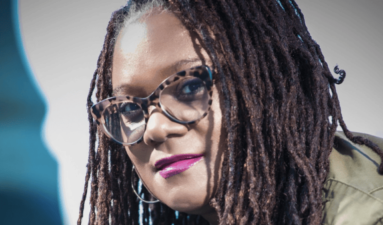 A woman with tortoiseshell glasses, braids, and purple lipstick wearing a green jacket