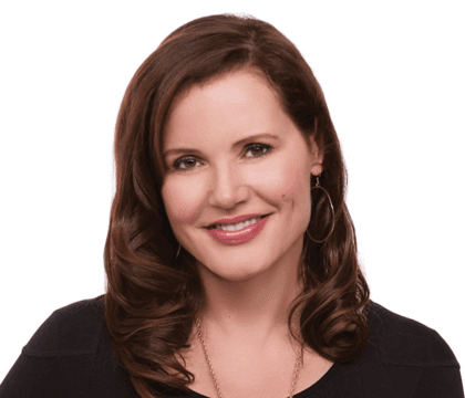 Geena Davis, wearing black top and gold necklace, smiles at camera